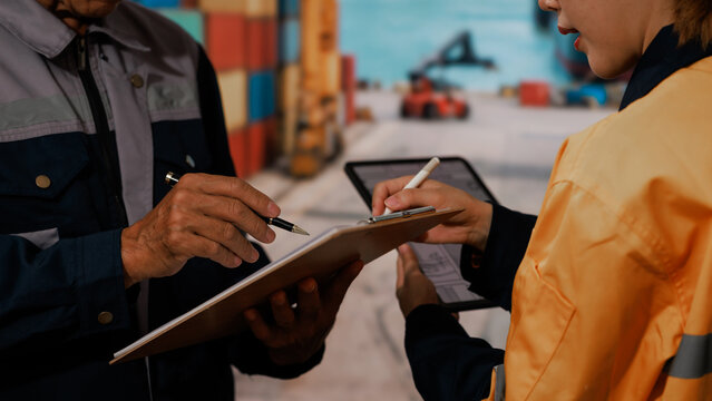 Warehouse workers review a clipboard and tablet while checking inventory in a busy loading area with containers and forklift equipment.