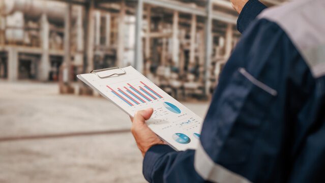 Factory worker in protective uniform reviews production charts on a clipboard inside an industrial warehouse, quality control and workplace inspection.