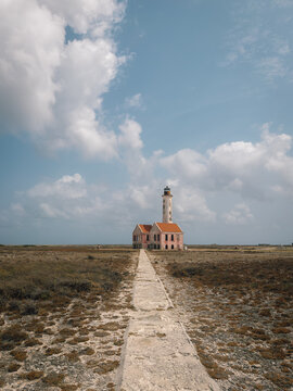 abandoned lighthouse stationary in deserted coastal area with red roof and pathway leading towards concrete and brick structure, serene and desolate mood, ground-level perspective looking.