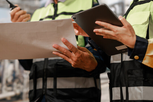 Close-up of construction workers in safety vests reviewing a blueprint and tablet at a work site, teamwork, planning and project management.