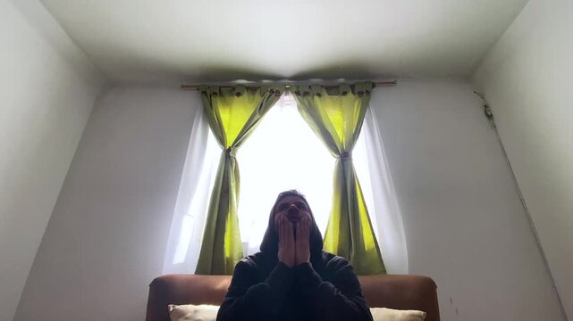Portrait of a young Colombian man expressing feelings of despair and frustration in front of two curtains in his room. Concept of mental health and emotional well-being 