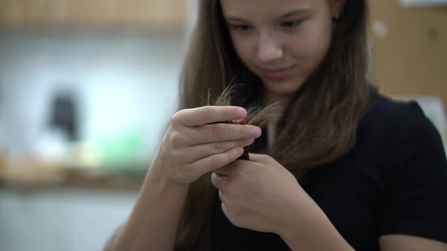 Young girl looks closely at the tips of a brown lock she holds, checking hair condition.