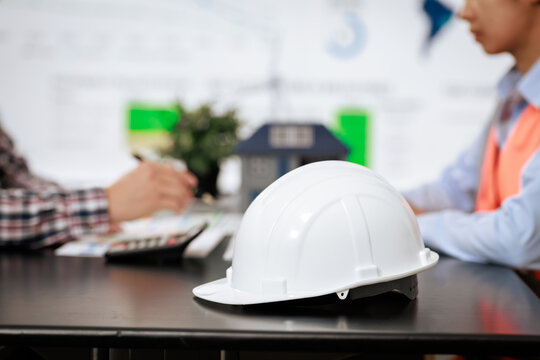 White safety helmet on an office desk during a meeting between construction professionals, with a small house model and paperwork in the background
