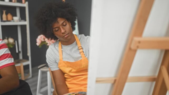 Two men painter friends in aprons, one holds paintbrush and palette while the other watches and gestures at canvas in studio; collaborative creativity.