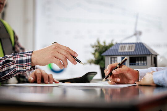 People signing property documents beside a model house with solar panels, a home purchase, real estate agreement, and renewable energy planning.