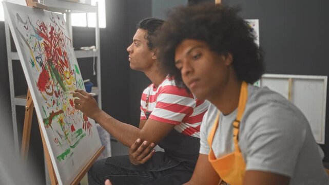Two men painters in aprons, one painting a canvas with a brush while the other sits watching and resting hands on lap in art studio; creativity collaboration.