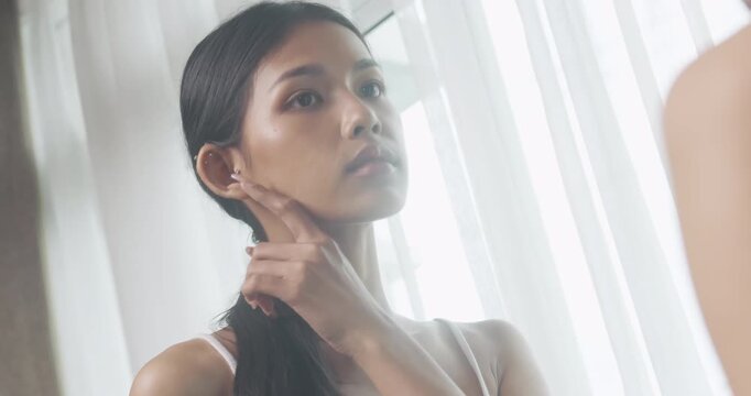 Young Asian woman touching her soft face skin in front of mirror. Beautiful girl checking clean skin and smooth texture at home. Daily morning skincare routine concept.