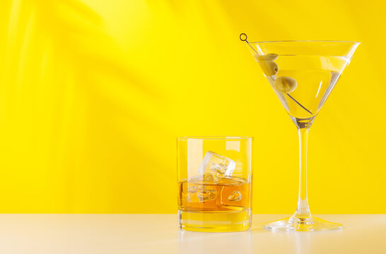 Glass of whiskey with ice and martini with olives on yellow background