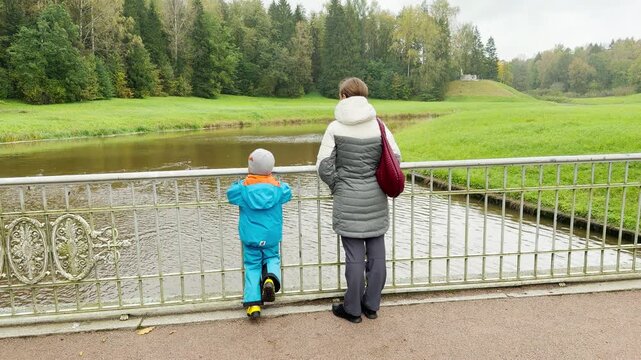 A mother and child stand on a bridge, looking at the river in Pavlovsk Park, Saint Petersburg. Green trees and hills surrounding them create a serene, happy, and peaceful atmosphere