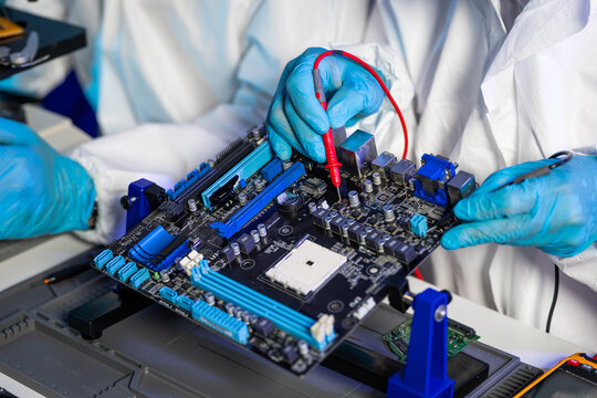 Technicians in protective suits and blue gloves test a computer motherboard with probes during electronics repair and quality control.