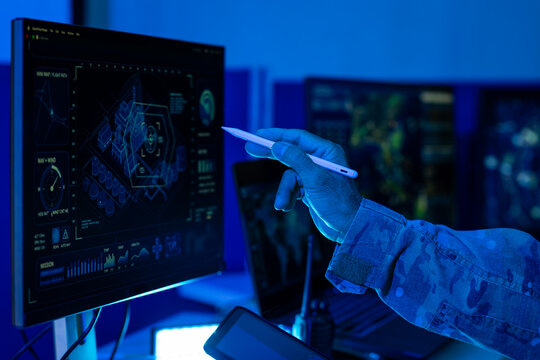 Military analyst in camouflage uniform uses a stylus on a touchscreen monitor digital security data in a blue command center.
