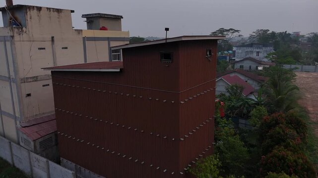 An aerial view shows a tall, brown building made of corrugated metal that serves as a swiftlet house, located next to a residential area filled with tropical trees under a cloudy sky.