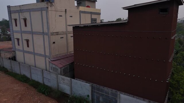 An aerial view shows a tall, brown building made of corrugated metal that serves as a swiftlet house, located next to a residential area filled with tropical trees under a cloudy sky.