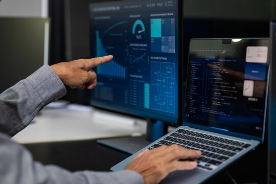 Close-up of a programmer using a laptop and pointing at a large monitor with data charts, code, and digital dashboard screens in a modern office.