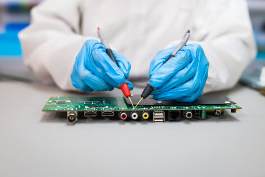 Technician in blue gloves testing an electronic circuit board with multimeter probes on a workbench.