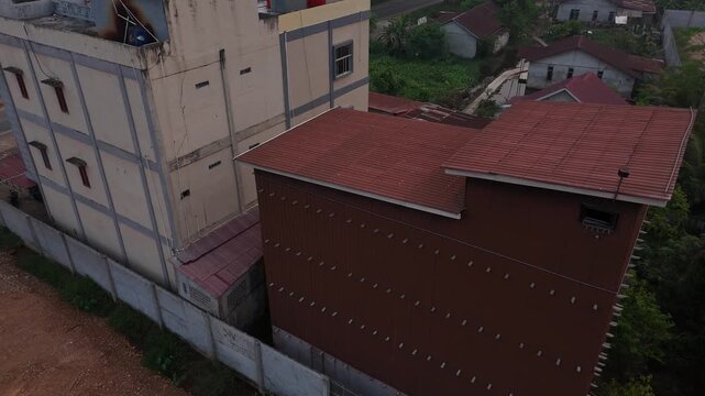 An aerial view shows a tall, brown building made of corrugated metal that serves as a swiftlet house, located next to a residential area filled with tropical trees under a cloudy sky.