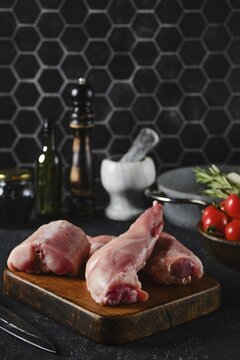 Rabbit hind legs sit on a wooden cutting board, surrounded by herbs, tomatoes, and kitchen tools. The sleek black hexagon tiles in the background create a stylish cooking atmosphere