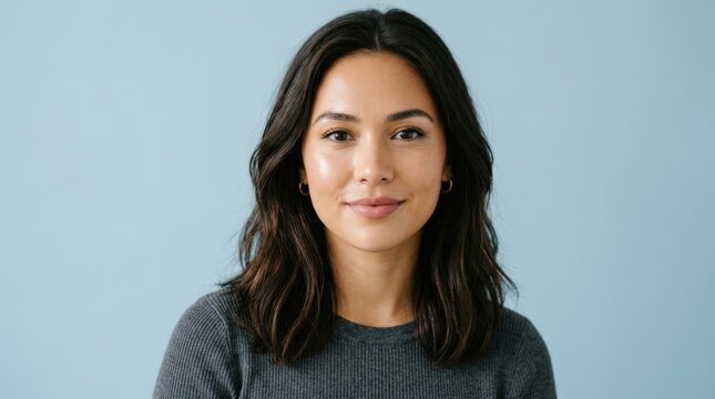 Partial portrait of a young woman with shoulder-length dark hair against a cool light-blue studio background, clean beauty photography, soft frontal lighting, natural expression and minimal