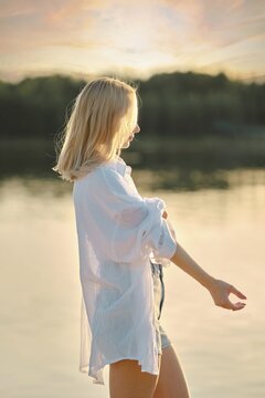 A young woman stands by the tranquil lake at sunset, wearing a white shirt and denim shorts. The soft sunlight casts a warm glow, highlighting her relaxed demeanor against the serene water backdrop