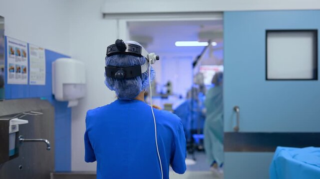 Following a female doctor wearing blue uniform and a cap with surgical loupes on. Surgeon walks to the surgery room approaching the operating table and waving her hands to dry them after washing.