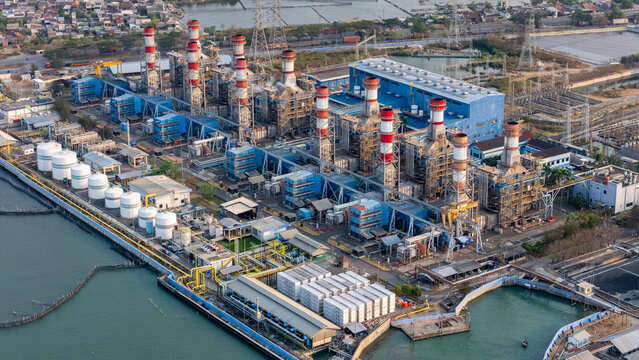 Aerial view of a large industrial power plant with tall smokestacks, numerous storage tanks, and complex infrastructure situated along the coast in Semarang, Central Java, Indonesia.