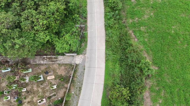 A drone shot from above showing a narrow concrete road separating a stretch of lush green fields and dense trees from a small cemetery with clearly visible headstones.