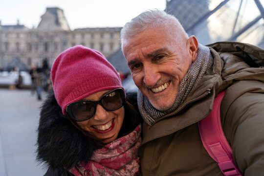 Smiling couple enjoying travel to paris during winter, wearing warm clothing, taking selfie in front of louvre