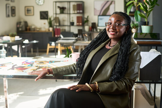 Portrait of smiling African American young adult woman sitting at desk in creative design studio. Image suitable for business branding, advertising and corporate marketing materials