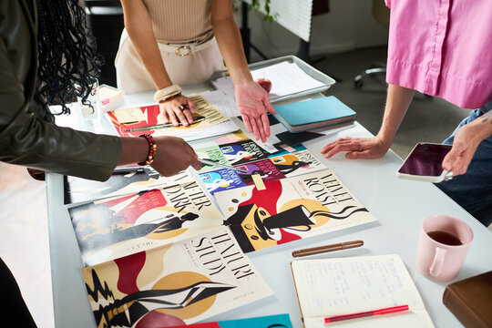 Multiethnic young adult women brand designers reviewing graphic posters during marketing meeting in creative studio. Scene suitable for corporate branding campaigns and teamwork concepts