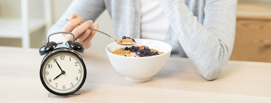 Young woman eating a healthy Greek yogurt bowl with granola and blueberries at a wooden table in the morning, with alarm clock showing the breakfast time.