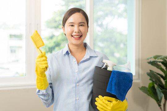 Cleaning professional housekeeping concept. Asian young woman wearing yellow gloves, hand holding dusting brush with tool bucket, smile girl looking at camera after doing housework or chores at home.