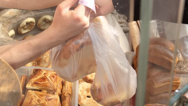 A bakery seller hands two packages of fresh bread to a customer through a service window and receives a banknote for the purchase. fresh bread, bakery trade, traditional bread, bread purchase