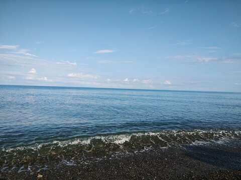 View of a blue sea under a light sky with small clouds. Gentle wave rolling onto a dark pebble beach. Coastal landscape background