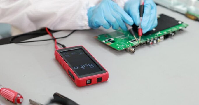 Technician in blue gloves tests an electronic circuit board with probes and a digital multimeter on a workbench.