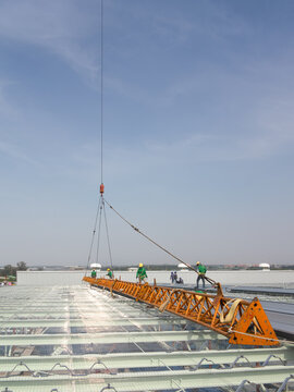 Crane hoists an orange steel lattice truss above a reinforced industrial floor as construction workers guide the load at Eastern Seaboard Industrial Estate in Rayong Thailand, during factory expansion