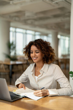 Vertical portrait of smiling latin business woman writing notes in notebook near laptop at coworking office workspace, planning. In 30s, female worker works as professional, businesswoman, manager