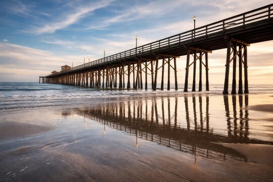 Long wooden pier over ocean at sunset with reflections on wet sand, peaceful coastal seascape and travel background.