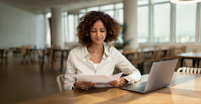Web banner of indian business woman reading paper document near laptop in coworking office workspace, checking paperwork. In 30s, female specialist works as consultant, director, founder. Copy space