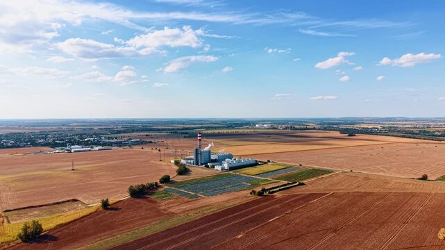 Farmland surrounding an industrial plant and solar installation. An aerial panorama shows a chimney, dark panel rows and crop plots beneath scattered clouds.