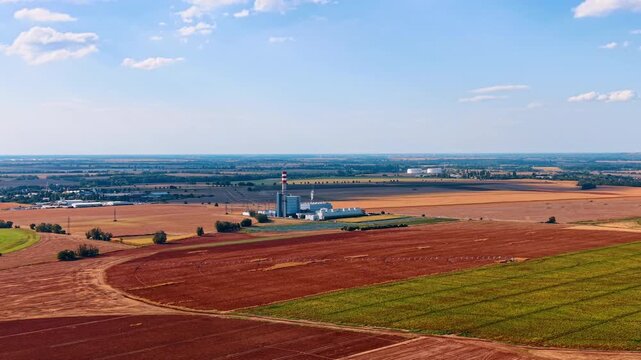 Industrial facility and solar array among cultivated fields. A striped chimney rises from buildings surrounded by brown farmland and rows of dark panels.