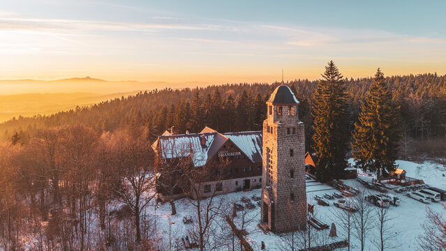 Bramberk lookout tower stands in winter with snow covering the ground. The tower is surrounded by trees and a building nearby. It is a quiet time in the late afternoon.