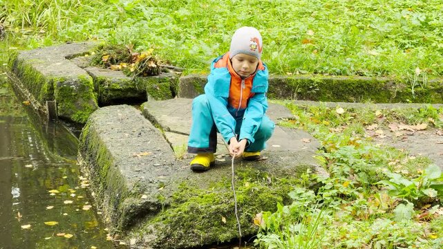 A young boy crouches on mossy stones, curiously dipping a stick into the quiet water. His playful discovery creates a feeling of joy in Pavlovsk Park, Saint Petersburg.