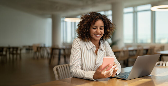 Web banner of smiling african business woman holding mobile phone near laptop in coworking office workspace, using app. In 30s, female boss works as leader, specialist, consultant. Copy space