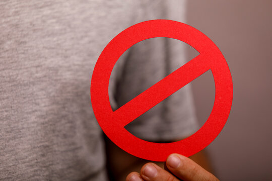 A person's hand holds a bright red circular prohibition symbol against a blurred background.