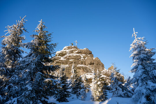 The cross sits on the summit of a rock formation in Jizera Mountain. Snow covers the trees and ground, creating a winter scene against the clear blue sky.
