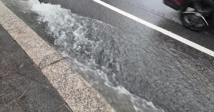Slow motion capture of a torrent of rainwater rushing along the sidewalk during a downpour, while vehicles drive by nearby.