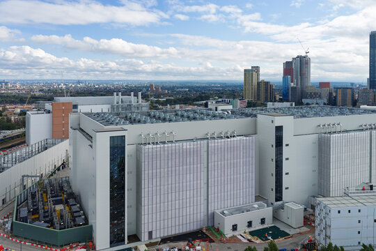 Aerial view of a large modern data center facility with rooftop cooling units, surrounded by urban infrastructure and a city skyline under a blue sky in the United Kingdom.