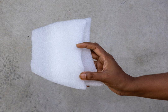 Close-up side view of a human hand holding a white poly foam sheet against gray concrete wall background.