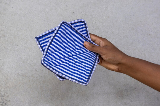 Close-up side view of human hand holding blue and white striped square quilted fabric pot holder against gray backdrop.