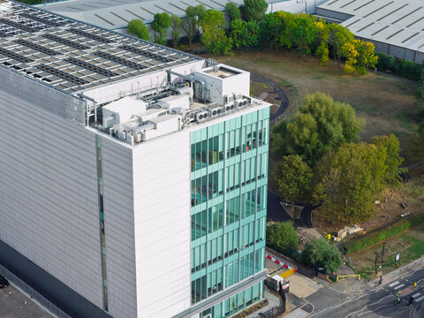 Aerial view of a modern data center with a glass facade and rooftop cooling units, surrounded by green trees and a road in the United Kingdom.